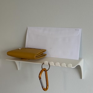 Könnte beinhalten: Ein weißes Wandregal mit einer gelben Geldbörse, einem weißen Umschlag und einem orangefarbenen Karabiner. Das Regal hat ein geschwungenes Design mit kleinen Haken. Die Geldbörse ist gefaltet und hat einen Reißverschluss.