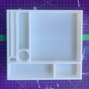 Puede incluir: Bandeja organizadora de plástico blanco con múltiples compartimentos de diferentes tamaños. La bandeja tiene un compartimento rectangular grande, un compartimento rectangular más pequeño, un compartimento circular y un compartimento largo y estrecho.