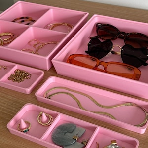 Puede incluir: Bandejas de plástico rosa con compartimentos para organizar joyas y gafas de sol. Las bandejas están llenas de varios collares, pulseras, pendientes y gafas de sol dorados.