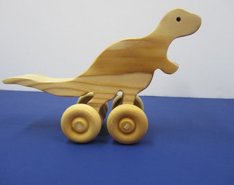 May include: A wooden toy dinosaur with four wheels. The dinosaur is light brown and has a long tail. The wheels are also wooden and are painted a light brown color.