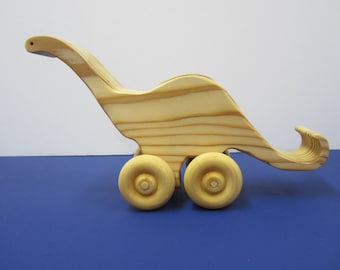 May include: A wooden toy dinosaur with a long neck and tail, painted in a natural wood color. The dinosaur has four wheels and is ready for playtime.