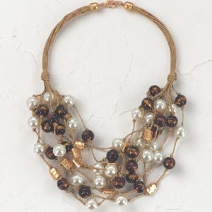 May include: A multi-strand necklace featuring tan cord strings adorned with white faux pearls, dark brown marbled beads, and small metallic gold-toned rectangular accents. The necklace is secured with a gold-toned lobster claw clasp closure.