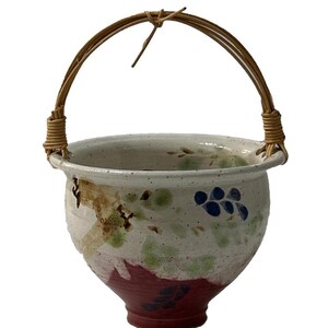 May include: A white ceramic planter with a red, green, and blue floral design. The planter has a woven bamboo handle.
