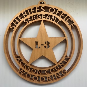 May include: A wooden sheriff's badge with a five-pointed star in the centre. The badge is inscribed with "Sheriff's Office Sergeant Jackson County Woodring" and "L-3".