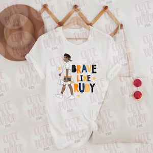 May include: White t-shirt featuring a graphic of a girl walking with a book, and the text "BRAVE LIKE RUBY" in blue, orange, and black. The shirt is hanging on a wooden hanger. A pair of red sunglasses is on the right.