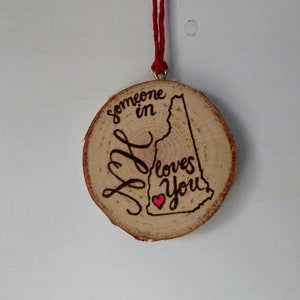 Pode incluir: Um ornamento de madeira em forma do estado de New Hampshire com o texto "Alguém em NH te ama" e um pequeno coração vermelho.