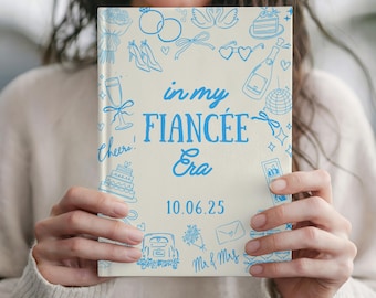 Op de afbeelding: Een crèmekleurig notitieboek met blauwe illustraties en tekst. De omslag leest "in my FIANCÉE Era" met de datum "10.06.25". Het notitieboek is versierd met tekeningen van huwelijksgerelateerde items zoals ringen, schoenen en taart.