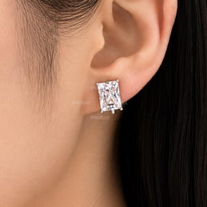 May include: A rectangular-cut clear gemstone stud earring set in a metallic prong setting. The earring is positioned on the earlobe, featuring a faceted surface that reflects light with a brilliant, transparent appearance and a secure metal backing.