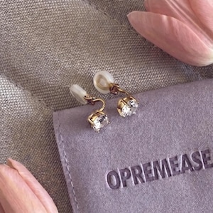 May include: A pair of gold-colored clip-on earrings with clear, square-cut gemstones. The earrings are displayed on a purple pouch with the word "OPREMEASE" printed on it, next to a pink tulip.