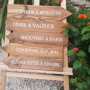May include: Wooden sign with white text pointing to different locations. The sign reads: Dancefloor &agrave; Beyrouth, Diner &agrave; Valence, Shooting &agrave; Paris, Cocktail &agrave; Dubai, Guinguette &agrave; Ein-Ebl.