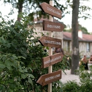 May include: Wooden signpost with five arrows pointing in different directions. Each arrow has a white text label: Cocktail &agrave; Dubai, Diner &agrave; Valence, Guinguette &agrave; Ein-Erl, Dancefloor &agrave; Beyrouth, Shooting &agrave; Paris.