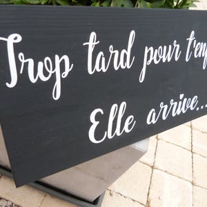May include: A black wooden sign with white lettering that reads "Trop tard pour t'enfuir Elle arrive..."