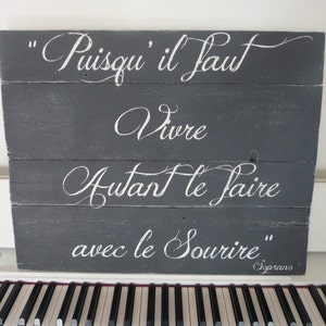 May include: A gray wooden sign with white lettering that reads "Puisqu'il faut vivre autant le faire avec le sourire" and "Soprano" in the bottom right corner.