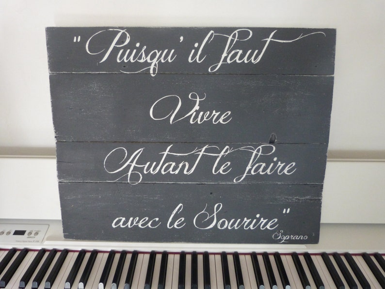 May include: A gray wooden sign with white lettering that reads "Puisqu'il faut vivre autant le faire avec le sourire" and "Soprano" in the bottom right corner.
