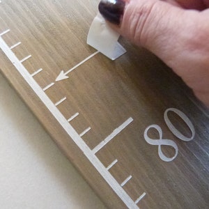 May include: Wooden height chart ruler with white markings and numbers. The chart is made of light brown wood and features a white arrow pointing to the 203 cm mark. A hand is peeling off a sticker.