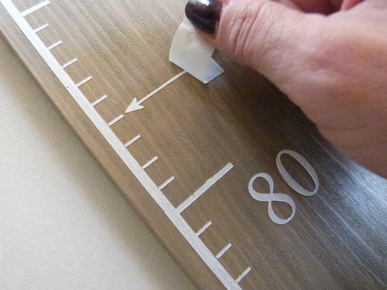 May include: Wooden height chart ruler with white markings and numbers. The chart is made of light brown wood and features a white arrow pointing to the 203 cm mark. A hand is peeling off a sticker.