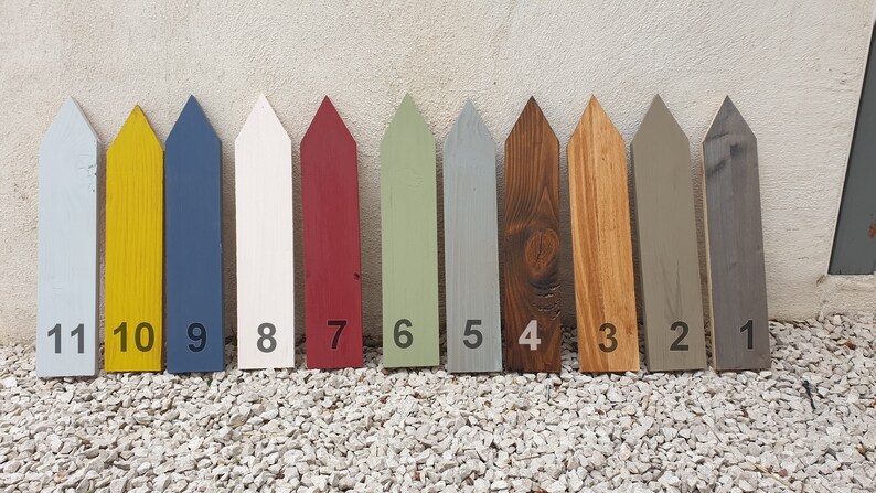 May include: Eleven painted wooden fence post markers in various colours. The colours include light blue, yellow, navy, white, red, green, grey, brown, tan, and dark grey. Each post is numbered from 1 to 11.