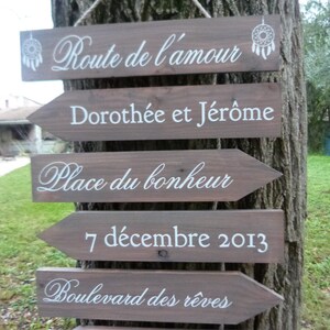 May include: A set of wooden signs with white text, hanging from a tree. The signs are shaped like arrows and point to the right. The text on the signs reads: "Route de l'amour", "Doroth&eacute;e et J&eacute;r&ocirc;me", "Place du bonheur", "7 d&eacute;cembre 2013", "Boulevard des r&ecirc;ves", and "Lola, Carmen, Swan".