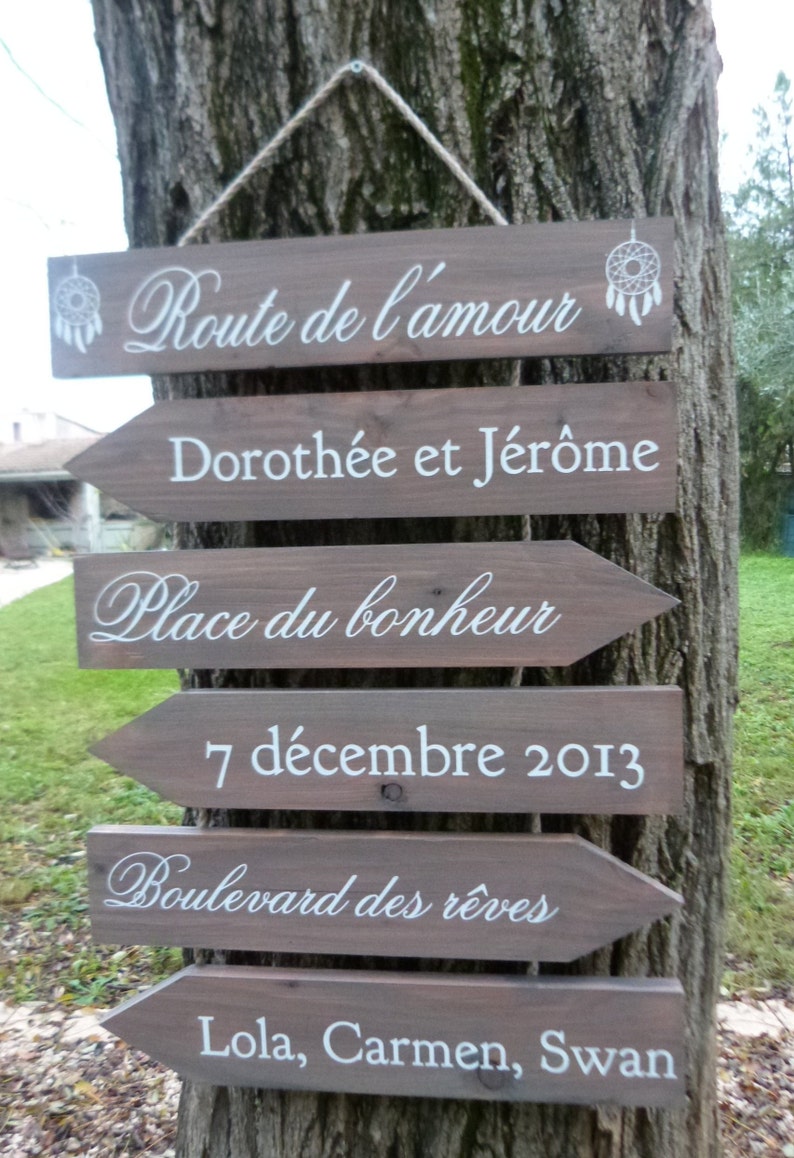 May include: A set of wooden signs with white text, hanging from a tree. The signs are shaped like arrows and point to the right. The text on the signs reads: "Route de l'amour", "Doroth&eacute;e et J&eacute;r&ocirc;me", "Place du bonheur", "7 d&eacute;cembre 2013", "Boulevard des r&ecirc;ves", and "Lola, Carmen, Swan".
