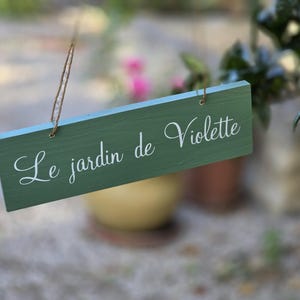Può includere: Una targa rettangolare in legno verde salvia appesa a un sottile spago marrone. La targa presenta la scritta in corsivo bianco Le jardin de Violette stampata al centro. Il legno ha una venatura visibile e una finitura liscia.