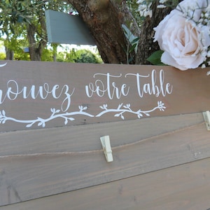 Peut inclure: Panneau en bois clair avec l'inscription "Trouvez votre table" en blanc. Décoration de vigne et corde avec pinces à linge. Des fleurs blanches sont visibles en arrière-plan.