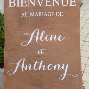 May include: A wooden sign with white text announcing the marriage of Aline and Anthony on 10 August 2019. The sign reads "Bienvenue au mariage de Aline et Anthony 10 ao&ucirc;t 2019".
