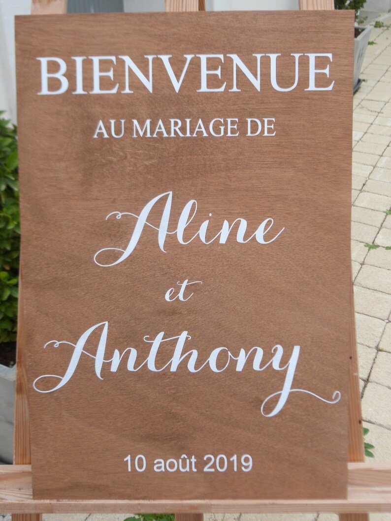 May include: A wooden sign with white text announcing the marriage of Aline and Anthony on 10 August 2019. The sign reads "Bienvenue au mariage de Aline et Anthony 10 ao&ucirc;t 2019".