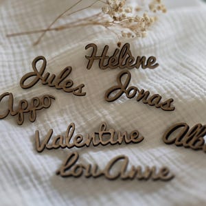 May include: A collection of laser-cut wooden name tags featuring the names Jules, Hélène, Appo, Jonas, Valentine, Alba, and Lou-Anne in a cursive script font, arranged on a white textured fabric surface.