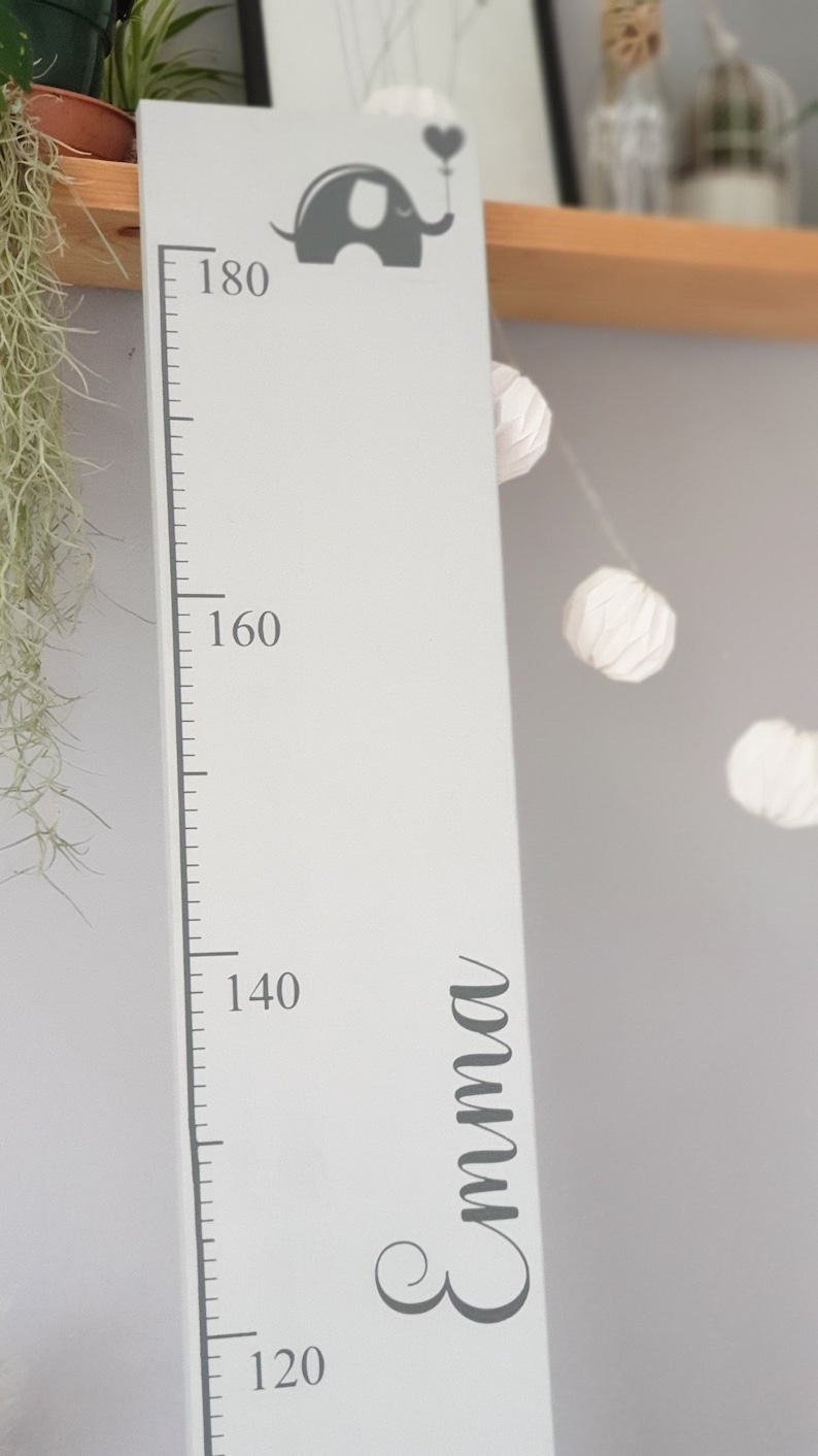 May include: A white wooden height chart with a grey elephant holding a heart-shaped balloon. The chart measures up to 180 cm and has the name "Emma" in cursive. A decorative item for a child's room.