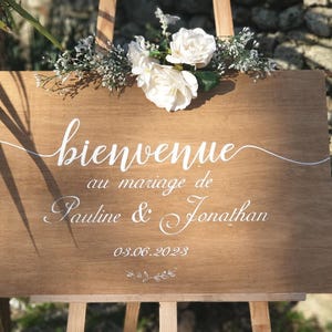 Puede incluir: Un letrero de madera con caligrafía blanca que dice "Bienvenue au mariage de Pauline & Jonathan 03.06.2023". El letrero está decorado con flores blancas y vegetación.