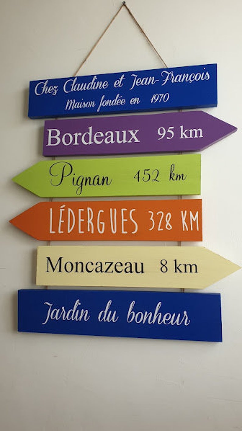 May include: A set of six wooden signs with arrow shapes, painted in different colors, with white text indicating distances to various locations. The top sign is blue and reads "Chez Claudine et Jean-Fran&ccedil;ois Maison fond&eacute;e en 1970". The signs below read "Bordeaux 95 km", "Pignan 452 km", "L&eacute;dergues 328 KM", "Moncazeau 8 km", and "Jardin du bonheur".