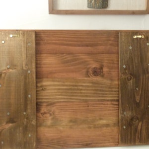 May include: The back of a wooden sign with two brown wood panels attached to the sides. The panels have several small silver screws holding them in place.
