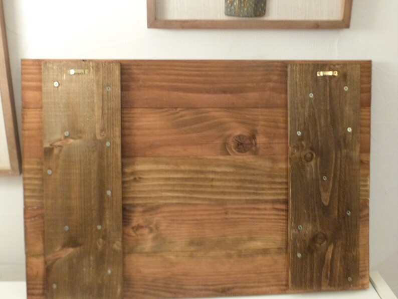 May include: The back of a wooden sign with two brown wood panels attached to the sides. The panels have several small silver screws holding them in place.