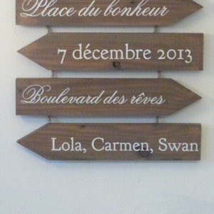 May include: Four wooden arrow signs with white text. The top sign reads "Place du bonheur". The second sign reads "7 d&eacute;cembre 2013". The third sign reads "Boulevard des r&ecirc;ves". The bottom sign reads "Lola, Carmen, Swan".