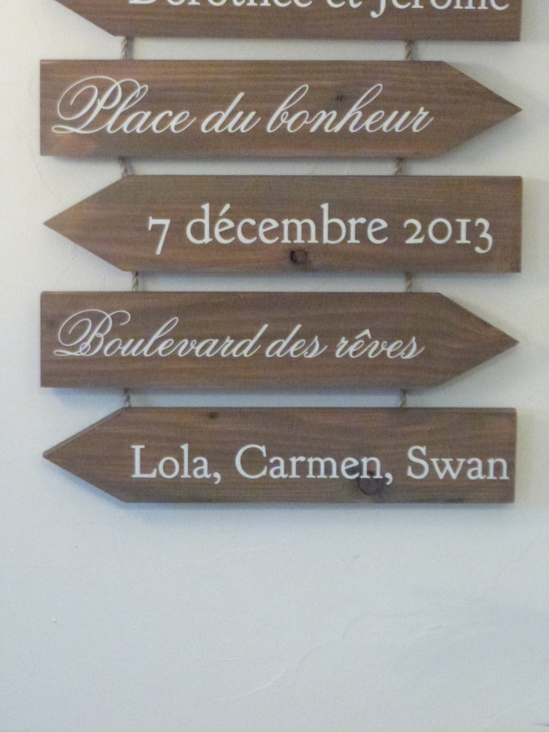 May include: Four wooden arrow signs with white text. The top sign reads "Place du bonheur". The second sign reads "7 d&eacute;cembre 2013". The third sign reads "Boulevard des r&ecirc;ves". The bottom sign reads "Lola, Carmen, Swan".