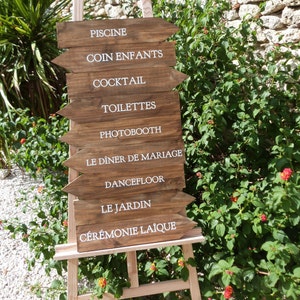 May include: Wooden sign with white text arrows pointing to different locations at a wedding. The sign reads: "Piscine", "Coin Enfants", "Cocktail", "Toilettes", "Photobooth", "Le Diner de Mariage", "Dancefloor", "Le Jardin", "Ceremonie Laique".