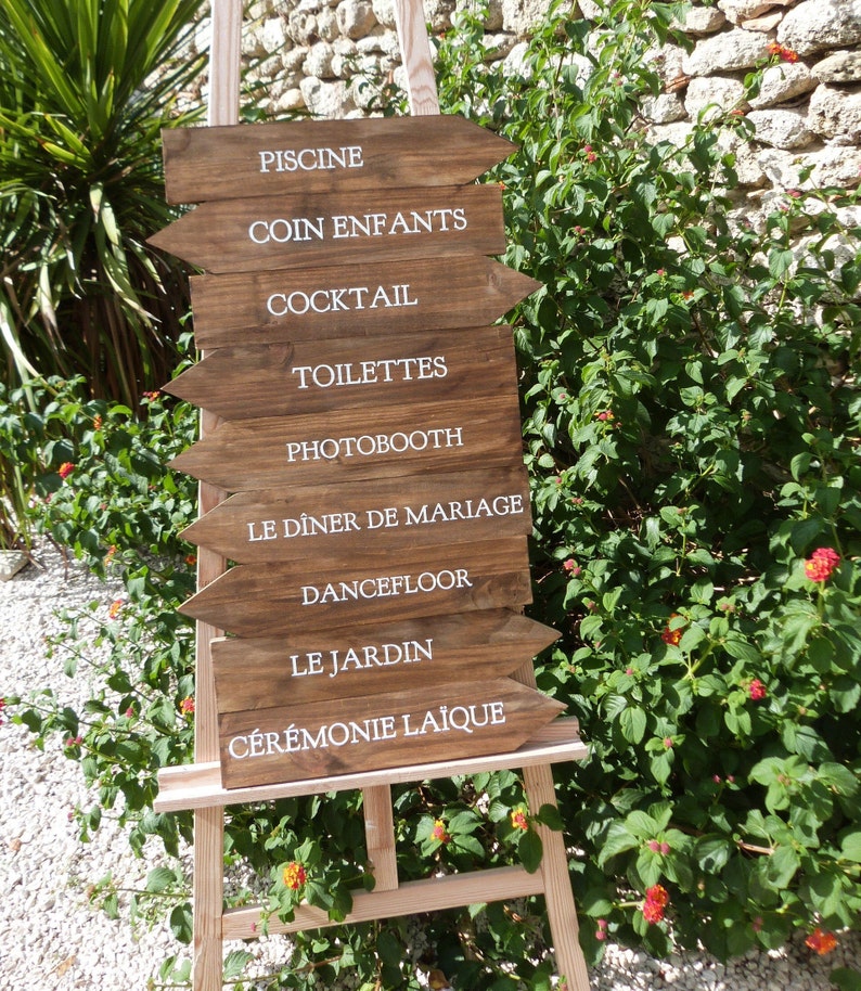May include: Wooden sign with white text arrows pointing to different locations at a wedding. The sign reads: "Piscine", "Coin Enfants", "Cocktail", "Toilettes", "Photobooth", "Le Diner de Mariage", "Dancefloor", "Le Jardin", "Ceremonie Laique".