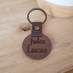 May include: A brown wooden keychain with the names "Jules Lucas" engraved on a round wooden pendant. The keychain has a leather strap and a brass-colored ring. The background is a light-colored wooden surface.