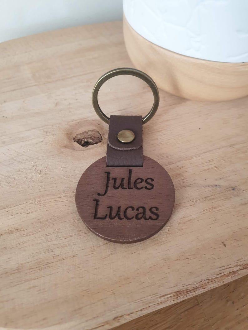 May include: A brown wooden keychain with the names "Jules Lucas" engraved on a round wooden pendant. The keychain has a leather strap and a brass-colored ring. The background is a light-colored wooden surface.