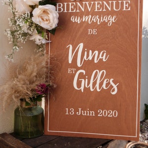 May include: A wooden sign with white text announcing a wedding for Nina and Gilles on 13 June 2020. The sign is decorated with white flowers and greenery.