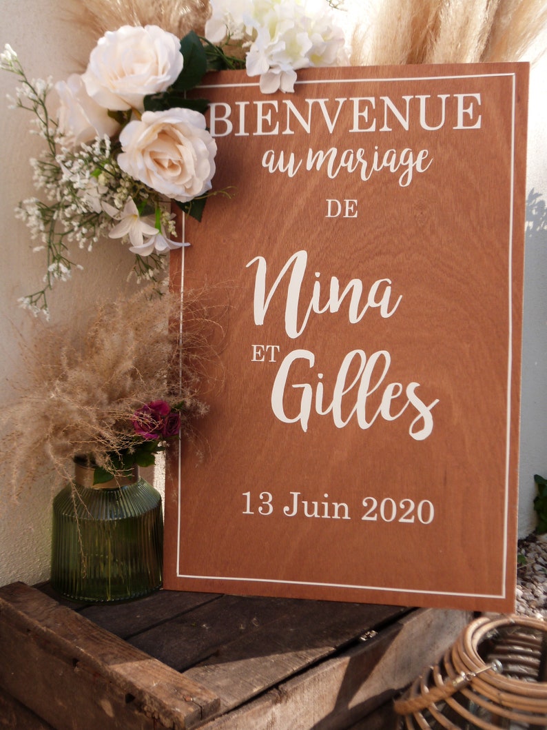 May include: A wooden sign with white text announcing a wedding for Nina and Gilles on 13 June 2020. The sign is decorated with white flowers and greenery.