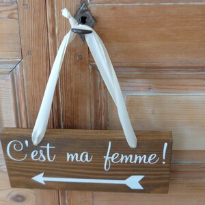 May include: A wooden sign with a white arrow pointing to the right and the text "C'est ma femme!" in white lettering. The sign is hanging from a white ribbon.