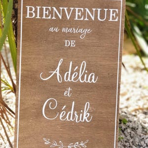 May include: A wooden wedding welcome sign with white text and a white border. The sign reads "Bienvenue au mariage de Adélia et Cédrik." A small floral design is at the bottom. The sign is set against a natural background.