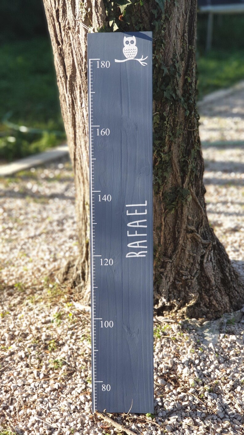 May include: A dark grey wooden height chart with white markings and the name "RAFAEL". An owl illustration is at the top. The chart measures up to 180 cm. The wooden plank is placed next to a tree.