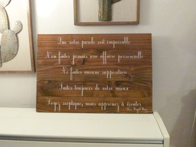 May include: A wooden sign with a white inscription in French. The text reads: "Que votre parole soit impeccable. N'en faites jamais une affaire personnelle. Ne faites aucune supposition. Faites toujours de votre mieux. Soyez sceptique, mais apprenez &agrave; &eacute;couter." The sign is on a white surface.
