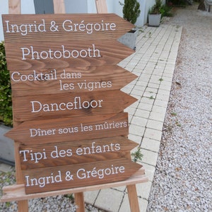 May include: A wooden sign with white text pointing to different activities at a wedding reception. The sign reads: Ingrid & Gr&eacute;goire, Photobooth, Cocktail dans les vignes, Dancefloor, D&icirc;ner sous les m&ucirc;riers, Tipi des enfants, Ingrid & Gr&eacute;goire.
