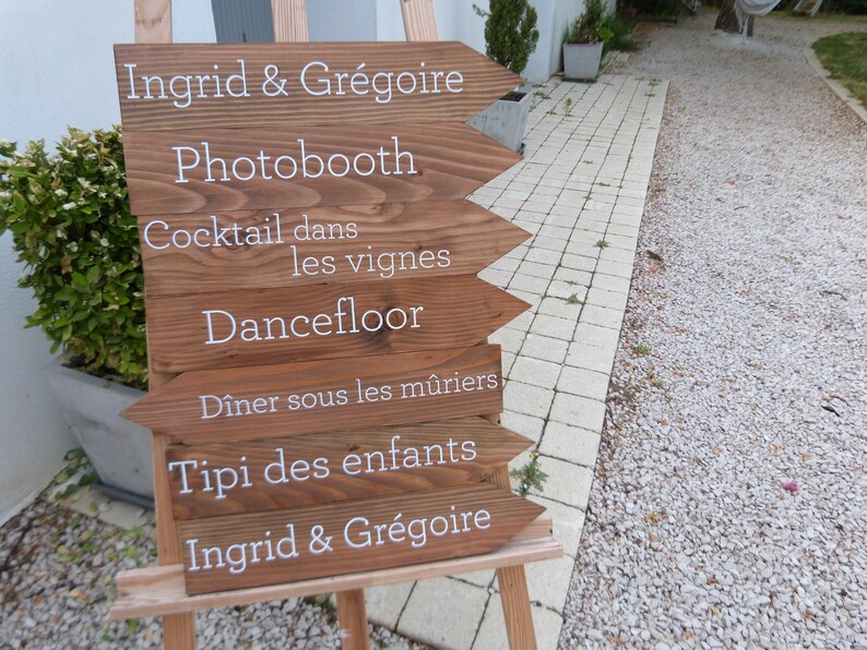 May include: A wooden sign with white text pointing to different activities at a wedding reception. The sign reads: Ingrid & Gr&eacute;goire, Photobooth, Cocktail dans les vignes, Dancefloor, D&icirc;ner sous les m&ucirc;riers, Tipi des enfants, Ingrid & Gr&eacute;goire.