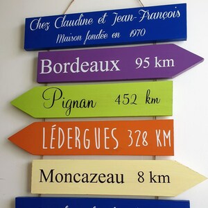 May include: A set of six wooden signs with arrow shapes, painted in different colors, with white text. The signs read: Chez Claudine et Jean-Fran&ccedil;ois Maison fond&eacute;e en 1970, Bordeaux 95 km, Pignan 452 km, L&Eacute;DERGUES 328 KM, Moncazeau 8 km, Jardin du bonheur.