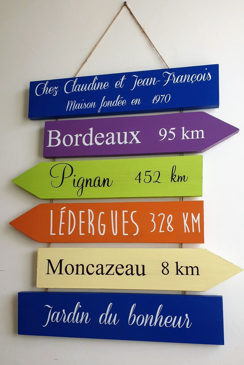 May include: A set of six wooden signs with arrow shapes, painted in different colors, with white text. The signs read: Chez Claudine et Jean-Fran&ccedil;ois Maison fond&eacute;e en 1970, Bordeaux 95 km, Pignan 452 km, L&Eacute;DERGUES 328 KM, Moncazeau 8 km, Jardin du bonheur.