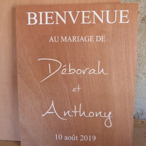 May include: A wooden sign with the text "BIENVENUE AU MARIAGE DE D&eacute;borah et Anthony 10 ao&ucirc;t 2019" in white lettering.